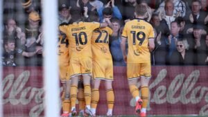 Five Newport County players in all amber celebrate scoring with a huddle in front of the crowd