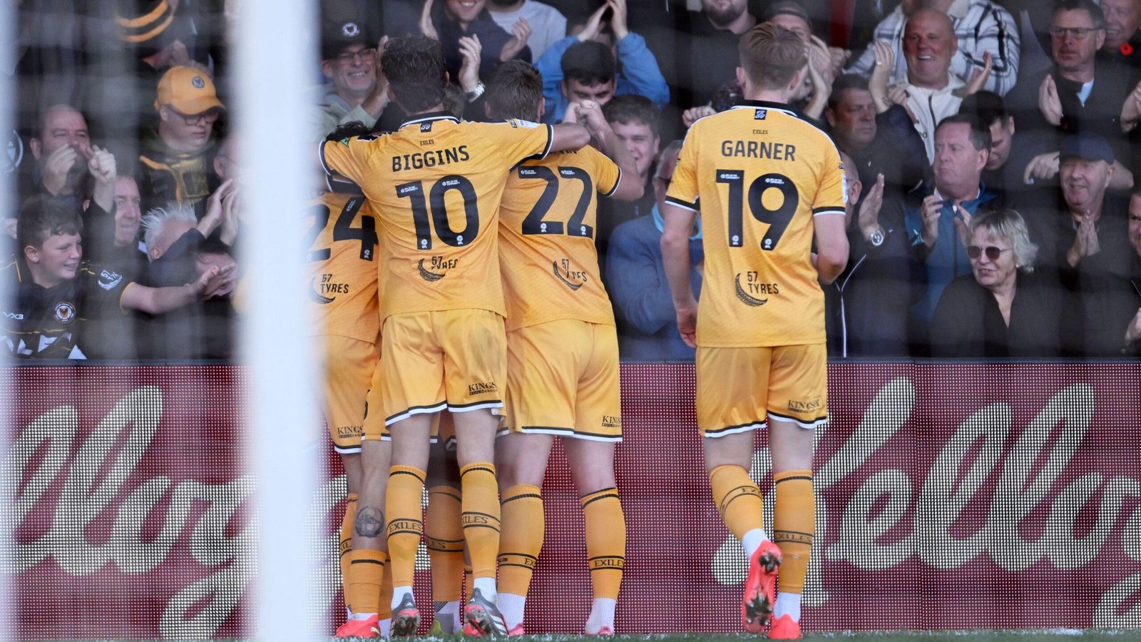 Five Newport County players in all amber celebrate scoring with a huddle in front of the crowd